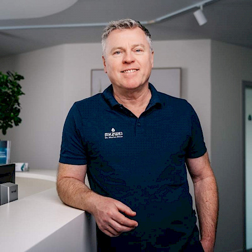 Porträt von Dr. Marius Klein, Zahnarzt in der Praxis MK2WEI in Dettingen an der Iller, lächelnd in blauem Poloshirt vor Praxisempfang.